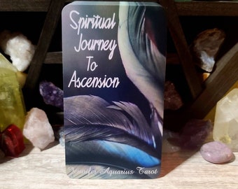 May include: A tarot card with a blue, gray, and white feather design. The text on the card reads "Spiritual Journey To Ascension" and "Jennifer Aquarius Tarot".