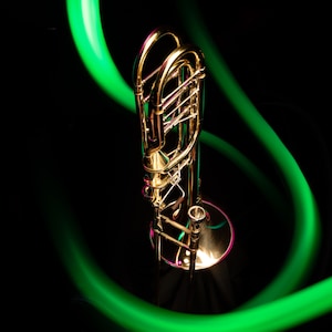 Könnte beinhalten: Ein goldfarbener Trombon mit einem grünen und violetten Lichtschweif dahinter. Der Trombon steht auf einer schwarzen Oberfläche.