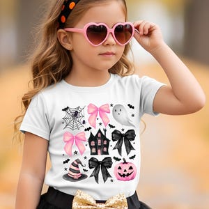 May include: A young girl in a Halloween costume, wearing a white t-shirt with Halloween graphics, a black tutu skirt, and striped leggings. She holds an orange jack-o'-lantern bucket and wears a polka-dot headband and heart-shaped sunglasses.