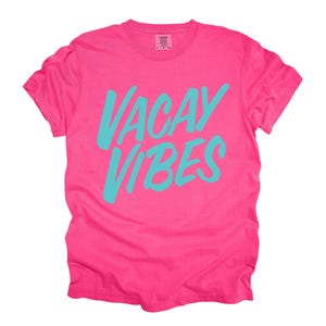 May include: A vibrant pink t-shirt with the words "VACAY VIBES" in turquoise script across the front. The shirt has a classic crew neck and short sleeves, perfect for a casual, summery look.