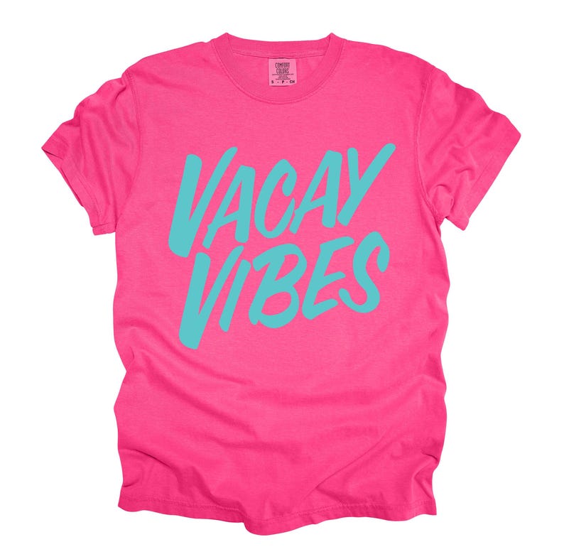 May include: A vibrant pink t-shirt with the words "VACAY VIBES" in turquoise script across the front. The shirt has a classic crew neck and short sleeves, perfect for a casual, summery look.