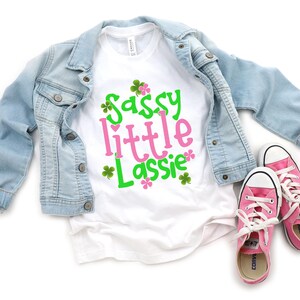 May include: White t-shirt with the text "Sassy little Lassie" in pink and green lettering with shamrock designs. The shirt is paired with a light blue denim jacket and pink sneakers.