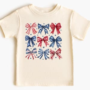 May include: A cream-colored t-shirt featuring a pattern of watercolor bows in red, white, and blue. The bows are arranged in a grid, with small blue stars scattered around them. The shirt is short-sleeved and has a crew neck.