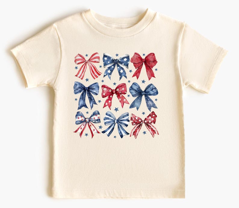 May include: A cream-colored t-shirt featuring a pattern of watercolor bows in red, white, and blue. The bows are arranged in a grid, with small blue stars scattered around them. The shirt is short-sleeved and has a crew neck.