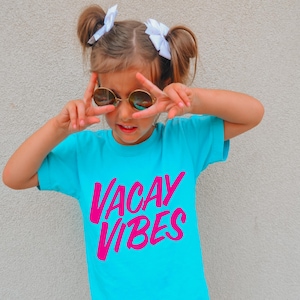 May include: A turquoise t-shirt with the words "VACAY VIBES" in bright pink. The person is wearing round sunglasses and making a peace sign with their fingers. The person has their hair up in two buns with white bows.