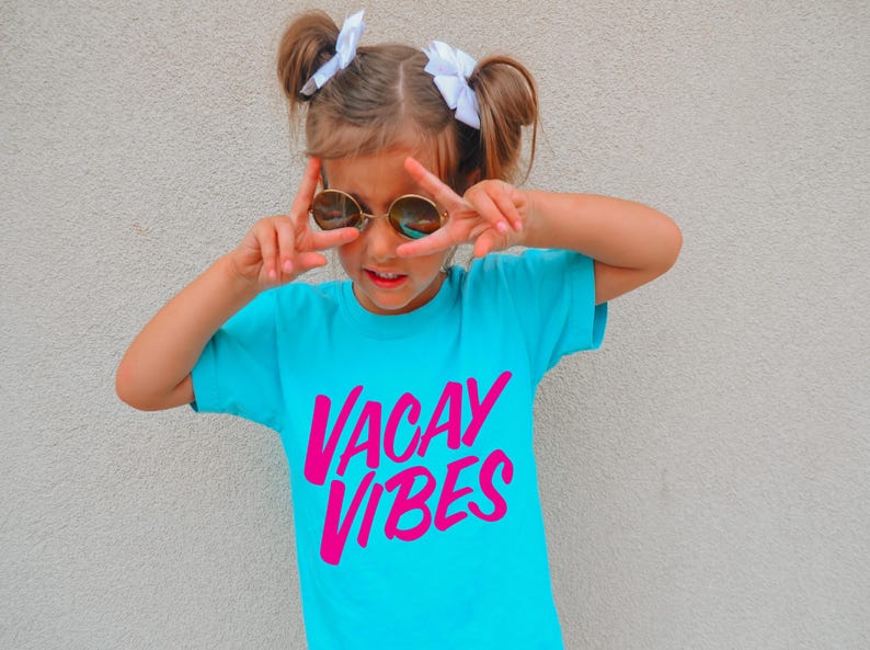 May include: A turquoise t-shirt with the words "VACAY VIBES" in bright pink. The person is wearing round sunglasses and making a peace sign with their fingers. The person has their hair up in two buns with white bows.