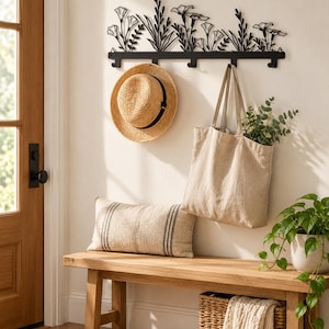May include: A black metal wall-mounted coat rack featuring a decorative silhouette of wildflowers and stems. The rack has five hooks holding a tan straw hat and a beige canvas tote bag filled with green foliage.