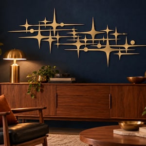 May include: A large, horizontal wall art piece made of gold-toned metal, featuring a cluster of multi-pointed starburst shapes and small circular accents. The design consists of elongated, sharp rays radiating from central points in a geometric arrangement.