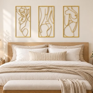 May include: Three rectangular wall art pieces featuring gold-toned wire outlines of human figures. Each piece is framed in a thin gold border, displaying continuous line art of abstract bodies mounted horizontally above a wooden bed frame.
