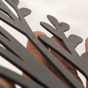 May include: A matte black, flat, laser-cut decorative object featuring a stylized, branching, leaf-like or fern-like silhouette with multiple rounded protrusions extending from a central stem, showcasing a textured surface finish.