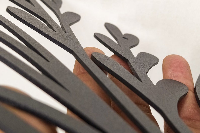 May include: A matte black, flat, laser-cut decorative object featuring a stylized, branching, leaf-like or fern-like silhouette with multiple rounded protrusions extending from a central stem, showcasing a textured surface finish.