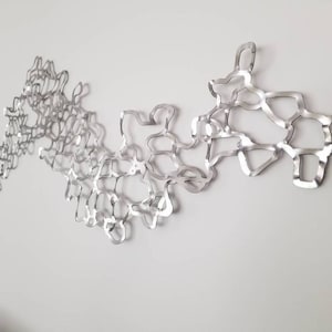 Könnte beinhalten: Eine silberne Wandskulptur aus Metall mit einem fließenden, organischen Design. Die Skulptur besteht aus miteinander verbundenen, wellenförmigen Formen, die ein Gefühl von Bewegung erzeugen.