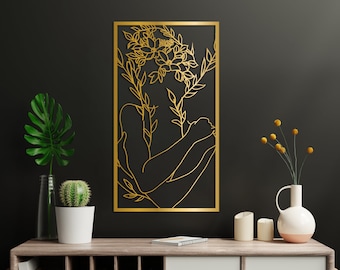 May include: Gold metal wall art featuring a couple embracing with floral accents. The artwork is framed in a thin gold frame and is mounted on a black wall.