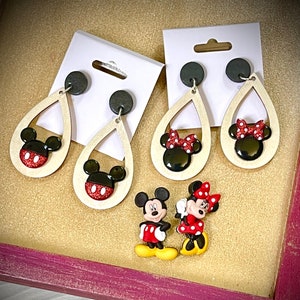 May include: A set of three pairs of dangle earrings featuring Mickey Mouse and Minnie Mouse. The earrings are made of wood and have a teardrop shape. The Mickey Mouse earrings have a black head with red and white polka dots and black ears. The Minnie Mouse earrings have a black head with red and white polka dots, black ears, and a red bow. The earrings are displayed on a wooden surface.