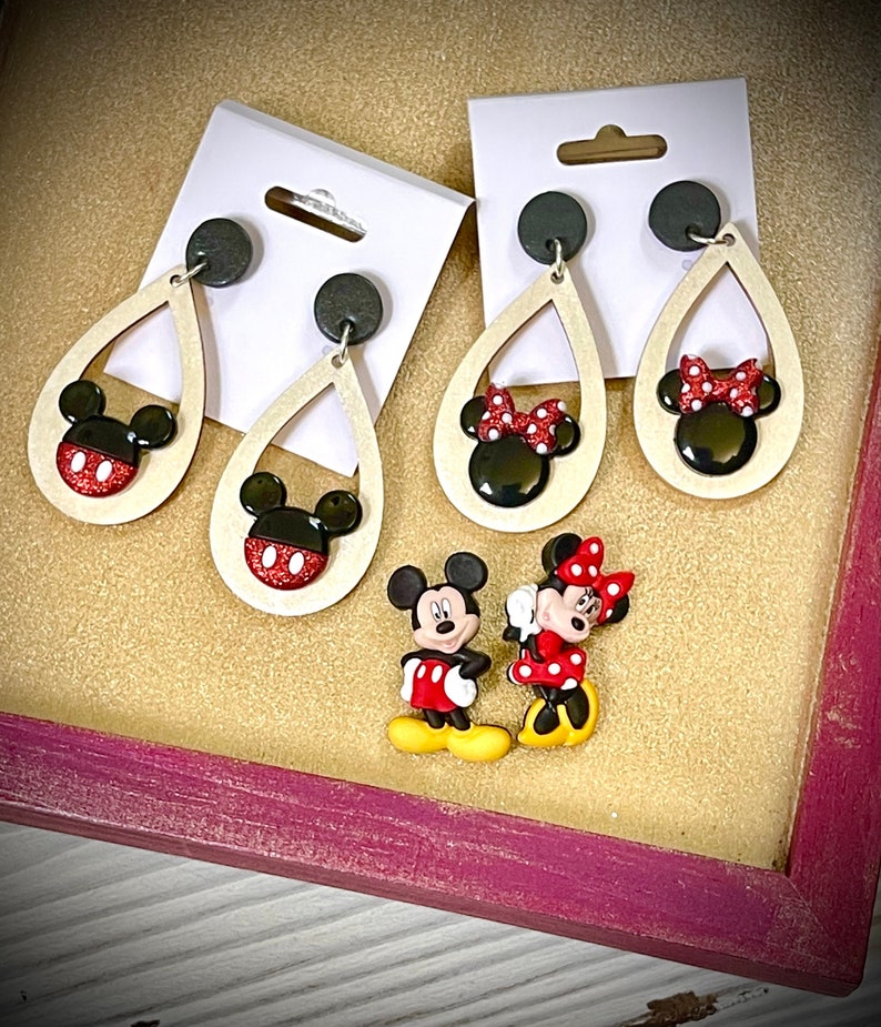 May include: A set of three pairs of dangle earrings featuring Mickey Mouse and Minnie Mouse. The earrings are made of wood and have a teardrop shape. The Mickey Mouse earrings have a black head with red and white polka dots and black ears. The Minnie Mouse earrings have a black head with red and white polka dots, black ears, and a red bow. The earrings are displayed on a wooden surface.