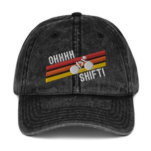Könnte beinhalten: Eine schwarze, vintage gewaschene Baseballkappe aus Denim mit einem weißen gestickten Design eines Radfahrers auf einem Fahrrad mit dem Text "OHHHH SHIFT!" im Retro-Stil.