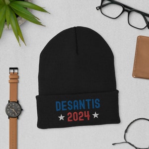 Puede incluir: Un gorro negro con la palabra "DESANTIS" en azul y "2024" en rojo, con estrellas blancas. El gorro está hecho de un material suave y es adecuado para el clima frío.