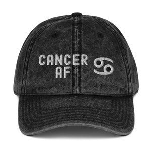 Könnte beinhalten: Verwaschene schwarze Baseballkappe mit dem gestickten Text "CANCER AF" und einem weißen Krebs-Tierkreiszeichen. Die Kappe hat einen gebogenen Schirm und einen Knopf oben.