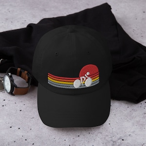 Puede incluir: Gorra de béisbol negra con un diseño bordado de estilo retro de un ciclista en una bicicleta frente a una puesta de sol. El diseño presenta una combinación de colores rojo, naranja, amarillo y gris.