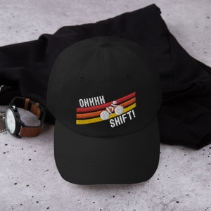 Könnte beinhalten: Schwarze Baseballkappe mit einem weißen gestickten Design eines Fahrrads mit dem Text "OHHHH SHIFT!" im Retro-Stil mit orangefarbenen, gelben und roten Streifen.