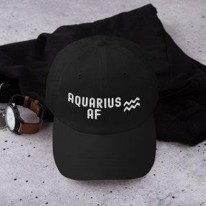 Könnte beinhalten: Schwarze Baseballkappe mit weiß gesticktem Text "Aquarius AF" und einem wellenförmigen Symbol.