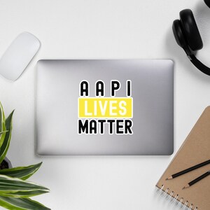 Puede incluir: Pegatina AAPI Lives Matter en un portátil plateado. La pegatina es texto negro sobre fondo amarillo.