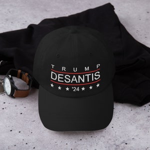 Puede incluir: Gorra de béisbol negra con el texto bordado "TRUMP DESANTIS '24" en blanco y rojo. La gorra tiene una visera curva y un diseño clásico de seis paneles. El texto está centrado en la parte delantera de la gorra, con estrellas arriba y abajo.