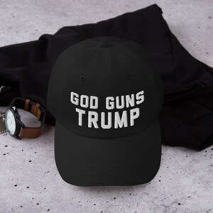 May include: Black baseball cap with white embroidered text that reads "GOD GUNS TRUMP".