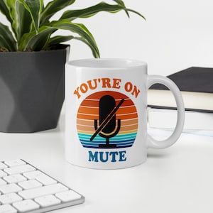 May include: White ceramic mug with a retro sunset design featuring a microphone with a line through it and the text "YOU'RE ON MUTE".