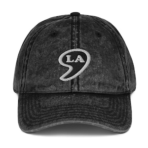 Puede incluir: Una gorra de béisbol negra de estilo vintage con un LA bordado en blanco dentro de una forma de coma.
