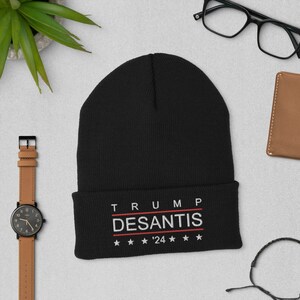 Puede incluir: Gorro de punto negro con el texto bordado en blanco "TRUMP DESANTIS '24" y cinco estrellas blancas a cada lado del año.