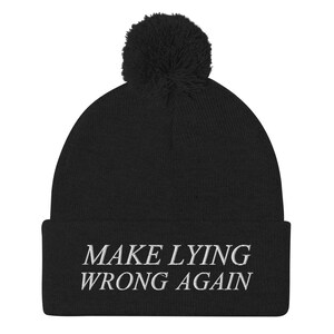 Może przedstawiać: Czarna czapka z dzianiny z białym pomponem na górze i haftem "MAKE LYING WRONG AGAIN" w kolorze białym na mankiecie.