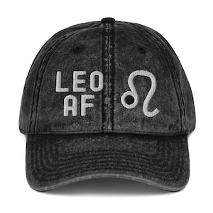 Könnte beinhalten: Schwarze, abgenutzte Baseballkappe aus Denim mit weiß gesticktem Text "Leo AF" und dem astrologischen Symbol für Löwe.