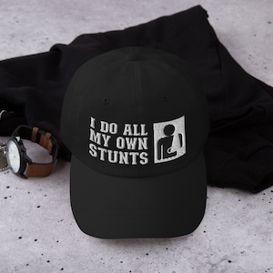 May include: Black baseball cap with white embroidery that reads "I do all my own stunts" and a stick figure with a broken arm.