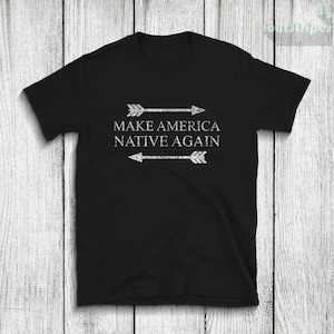 Op de afbeelding: Zwart T-shirt met witte tekst die "Make America Native Again" luidt met twee pijlen aan weerszijden van de tekst.