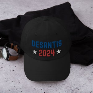 Puede incluir: Gorra de béisbol negra con el texto "DESANTIS 2024" bordado en azul y rojo con estrellas blancas a cada lado.