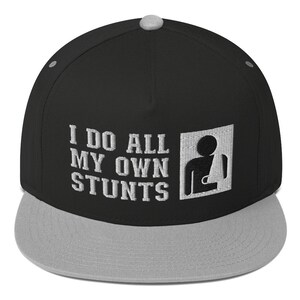 May include: Black and gray flat brimmed baseball cap with the text "I do all my own stunts" and a silhouette of a person with a broken arm.