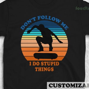 May include: Black t-shirt with a retro sunset graphic of a person skateboarding. The text "Don't Follow Me I Do Stupid Things" is printed on the shirt.