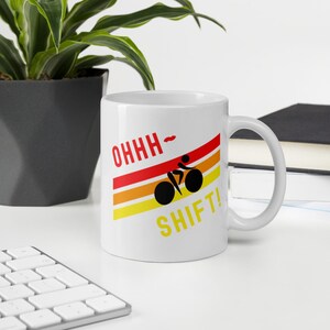 Könnte beinhalten: Weiße Keramiktasse mit einer schwarzen Silhouette eines Radfahrers auf einem Fahrrad, das einen Hügel hinauf fährt. Der Text "OHHH - SHIFT!" ist in roten, orangen und gelben Streifen auf die Tasse gedruckt.