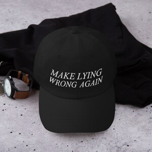 May include: A black baseball cap with white embroidered text that reads "MAKE LYING WRONG AGAIN".