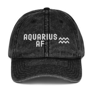 Könnte beinhalten: Schwarze, abgenutzte Baseballkappe aus Denim mit weiß gesticktem Text "Aquarius AF" und dem astrologischen Symbol des Wassermanns.