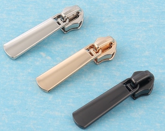 May include: Three zipper pulls in different finishes: silver, gold, and black. The pulls are rectangular with a rounded top and bottom.