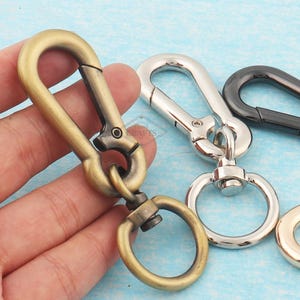 May include: Close-up of several metal carabiner clips in various colours. The primary clip is antique brass, connected to a ring. Other clips are silver and black. The background is a light blue surface.