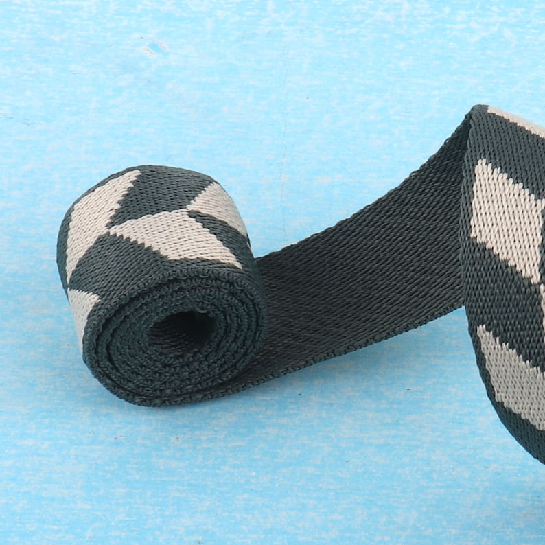 May include: A close-up of a rolled up dark green and white patterned webbing strap. The strap has a geometric pattern of white squares on a dark green background.