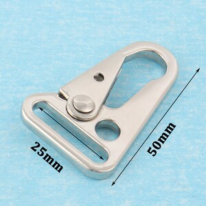 Puede incluir: Un cierre giratorio de metal plateado con un lazo rectangular de 25 mm y un gancho de 50 mm de largo. El cierre se muestra sobre un fondo azul claro.