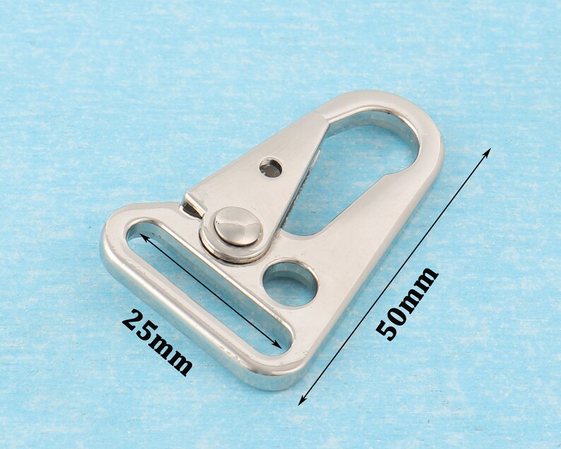 Puede incluir: Un cierre giratorio de metal plateado con un lazo rectangular de 25 mm y un gancho de 50 mm de largo. El cierre se muestra sobre un fondo azul claro.