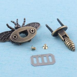 May include: A set of antique gold-toned metal hardware for a box or bag. The hardware includes a bee-shaped clasp, a spring-loaded catch, two screws, and a metal plate with five slots.