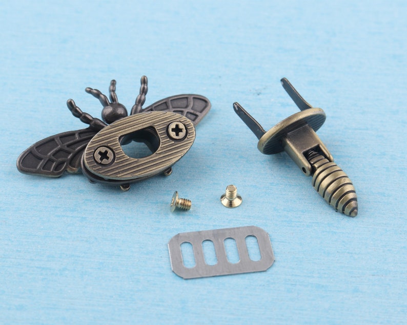May include: A set of antique gold-toned metal hardware for a box or bag. The hardware includes a bee-shaped clasp, a spring-loaded catch, two screws, and a metal plate with five slots.