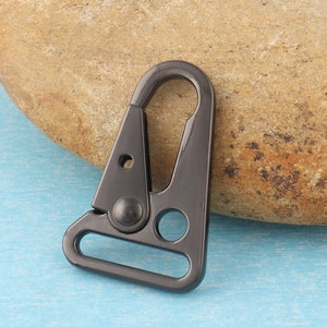 Puede incluir: Un cierre giratorio de metal gris oscuro con una puerta con resorte. El cierre tiene un cuerpo triangular con un lazo redondeado en la parte inferior y un agujero circular. El fondo es una superficie azul claro y una roca marr&oacute;n.