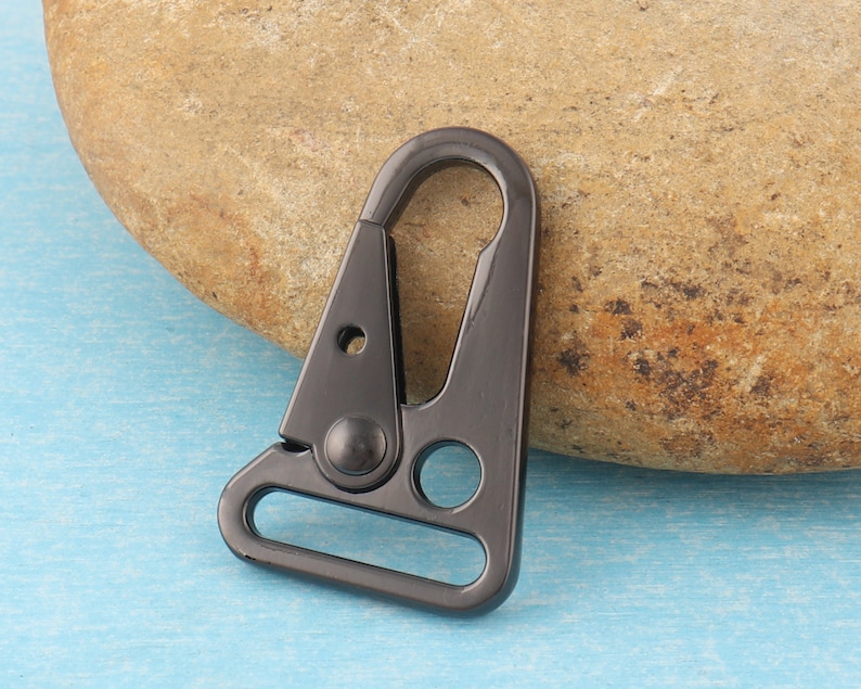 Puede incluir: Un cierre giratorio de metal gris oscuro con una puerta con resorte. El cierre tiene un cuerpo triangular con un lazo redondeado en la parte inferior y un agujero circular. El fondo es una superficie azul claro y una roca marr&oacute;n.
